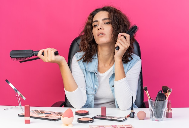 Confident pretty caucasian woman sitting at table with makeup tools holding combs and looking at side isolated on pink wall with copy space