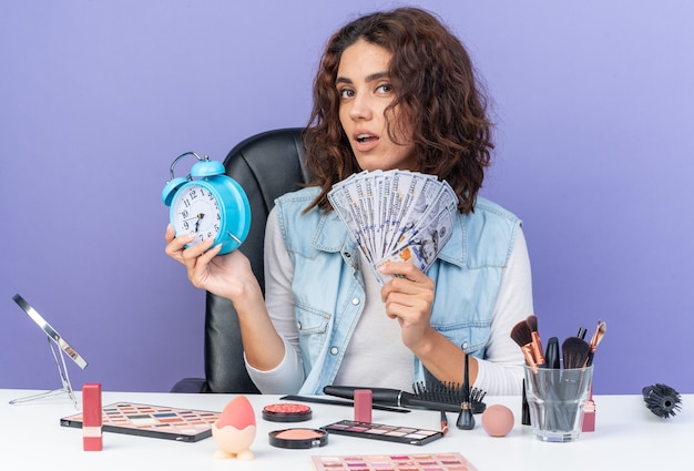 Confident pretty caucasian woman sitting at table with makeup tools holding money and alarm clock 