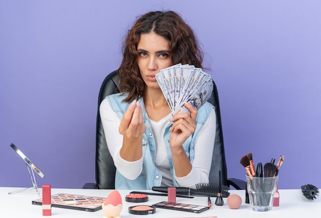 Confident pretty caucasian woman sitting at table with makeup tools holding money