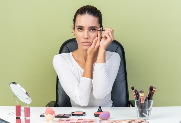 Donna abbastanza caucasica sicura che si siede al tavolo con strumenti per il trucco mettendo la mano sul viso e applicando l'eyeliner