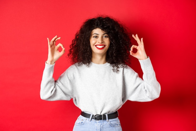 Confident pretty girl with curly hairstyle, showing okay gestures and smiling, approve and agree with you, praising excellent choice, standing satisfied on red wall