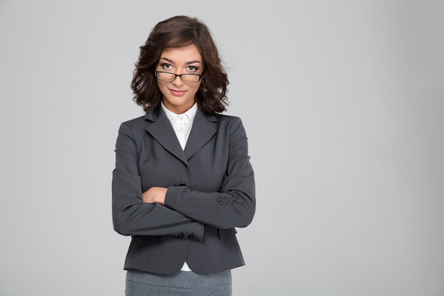 Confident pretty young curly businesswoman in glasses and gray jacket standing with crossed arms