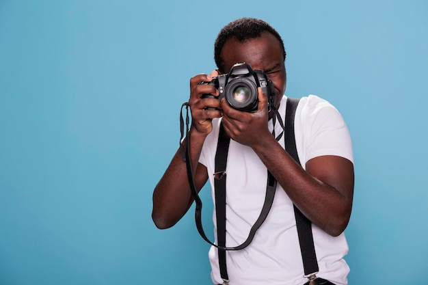 Fiducioso fotografo professionista con fotocamera dslr che scatta foto. appassionato di fotografia afroamericana che scatta foto stando in piedi su sfondo blu. studio girato