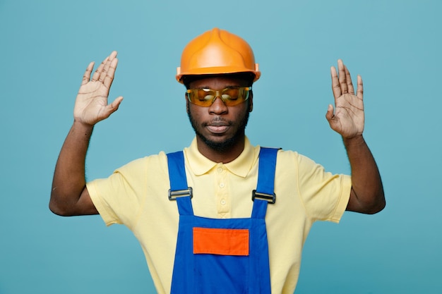 confident raisied hands young african american builder in uniform isolated on blue background