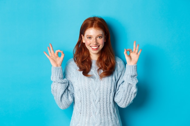 Confident redhead girl showing okay signs, praising choice, approve and agree, like something, standing over blue background