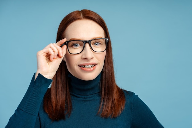 Confident redhead woman with braces smiling and looking at camera set against a blue background