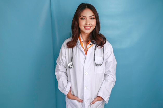 자신감이 있는 편안한 아시아 여성 의사가 실험복 주머니에 손을 넣고 파란색 배경 위에 복사 공간이 있는 카메라를 향해 조용히 웃고 있습니다.
