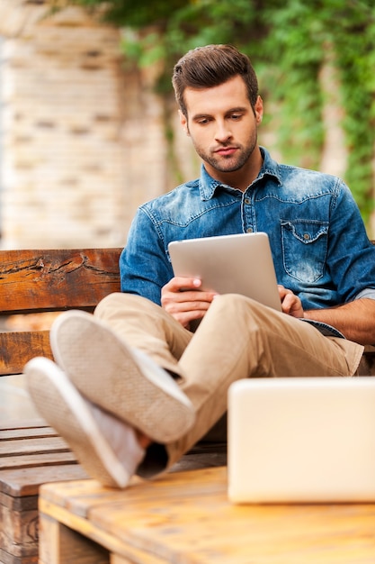 Confident and relaxed. Handsome young man working on digital tablet while sitting at the wooden table outdoors