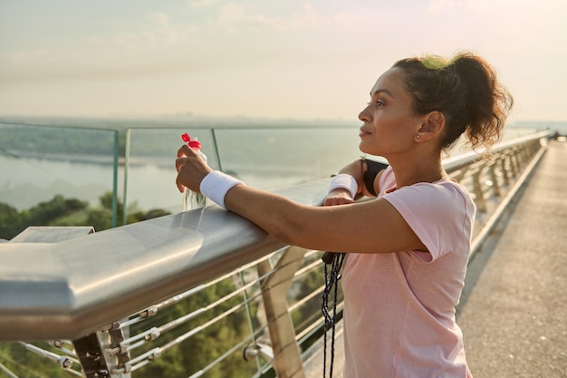 Confident relaxed middle aged sportswoman admiring beautiful nature standing on the city bridge after intense cardio workout at sunrise