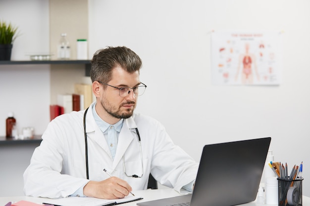 Confident senior doctor working with laptop at medical cabinet, bearded male therapist in white medical uniform and glasses sitting in med cabinet with stethoscope and using his personal computer