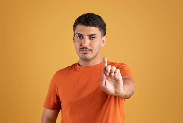 Confident serious brazilian man showing finger up stop sign standing isolated on orange background