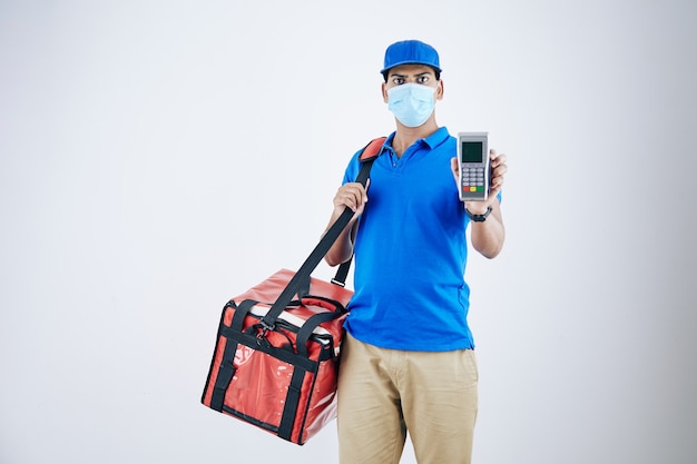 Confident serious delivery man in medical mask holding cooler bag showing payment terminal