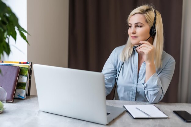 Confident serious experienced qualified beautiful smart woman with blonde hair wearing shirt is typing letters to her clients and business partners, sitting at the table in office