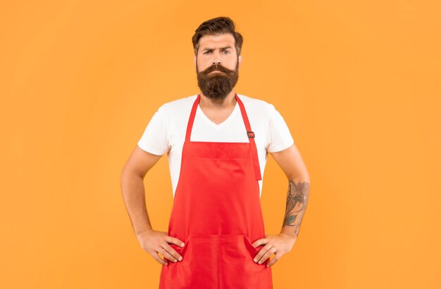 Confident serious man with arms akimbo in red apron yellow background household