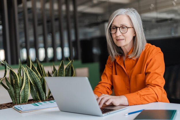 Уверенная в себе серьезная зрелая деловая женщина в стильных очках оранжевой рубашке с ноутбуком, работающая онлайн в современном офисе. Менеджер планирует проект, печатая на клавиатуре. Успешная деловая карьера.