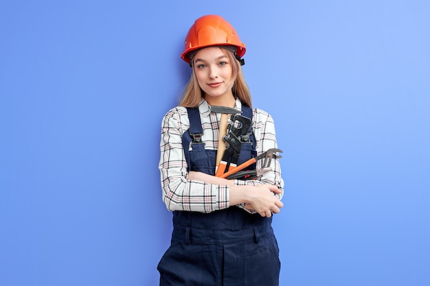 Confident service woman wearing orange helmet and blue coverall holding wrench, hammer and other tools in hands isolated over blue studio background