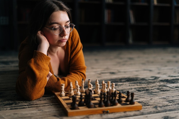 Confident sexy young woman in elegant eyeglasses thinking about chess move lying on wooden floor in dark room looking on chessboard