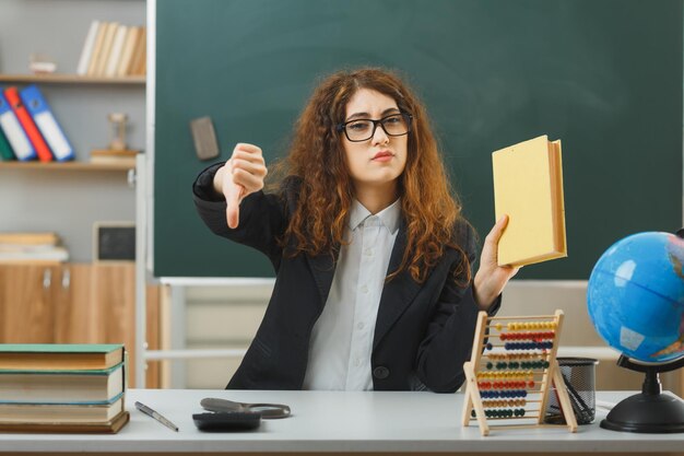 Fiducioso che mostra i pollici verso il basso giovane insegnante femminile che indossa occhiali tenendo il libro seduto alla scrivania con gli strumenti della scuola in classe