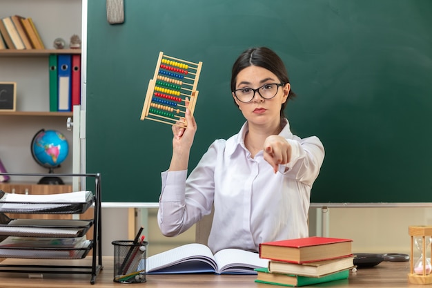 Fiducioso che ti mostra gesto giovane insegnante femminile che indossa occhiali con in mano l'abaco seduto al tavolo con gli strumenti della scuola in classe