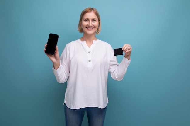 Confident slim blonde mature woman in white blouse holding plastic card for shopping with mockup