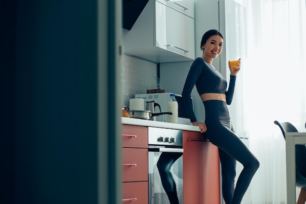 Confident slim lady in sports clothes standing by the cooker in her kitchen with a glass of juice Template banner