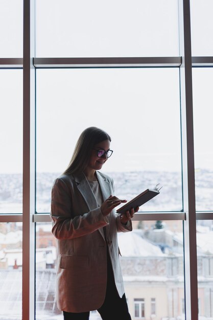 Confident slovenian business woman in elegant clothes checks notes in a notebook standing by the window in the office a serious female manager reads information viewed on a notebook