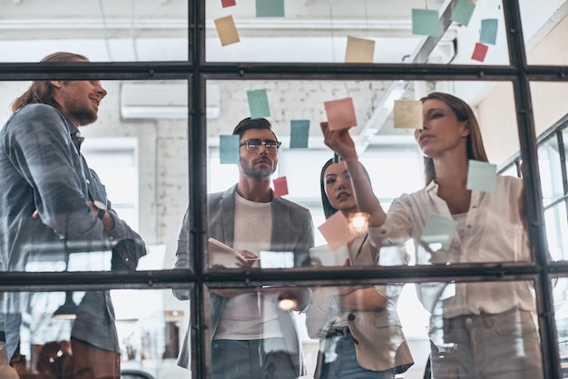 Confident and smart. Group of young modern people in smart casual wear using adhesive notes while standing behind the glass wall in the board room