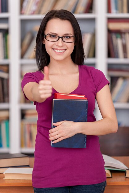 Confident and smart student. Cheerful young female student holding books and smiling while standing at the library