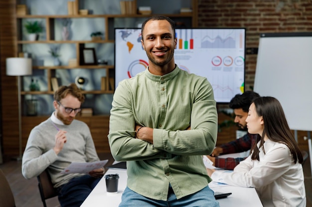 Confident smiling african american guy startup team leader stands against background of mixed race office workers or business people with arms crossed or clasped looking at the camera