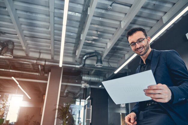 Confident smiling architect wearing in suit holding document and looking at it in the office