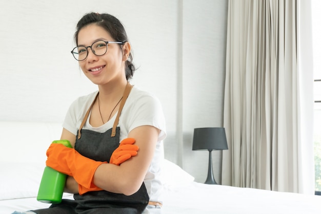 写真 灰色のエプロンで自信を持って笑顔のアジアの女性と彼女の寝室の掃除を準備するオレンジ色のゴム手袋。