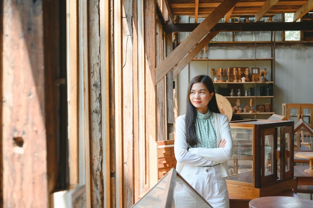 A confident smiling Asian woman stands near her desk at home and looks at the camera in a friendly manner She owns a piece of handcrafted wood Work in a wood crafts workshop