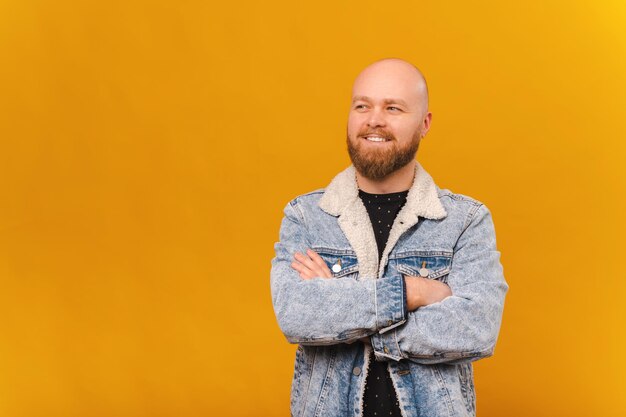 Confident smiling bearded bald man in jeans jacket is holding arms crossed