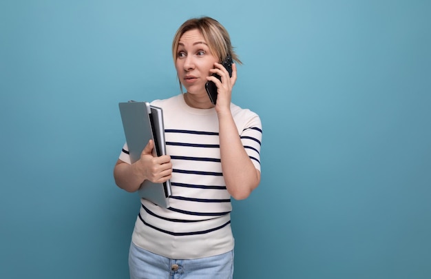 Confident smiling blond girl talking on a smartphone holding a laptop in her hands on a blue