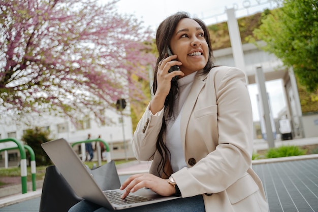 Donna sorridente sicura di affari che parla sul telefono cellulare usando il computer portatile che si siede nel parco