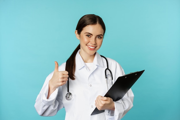 Confident smiling doctor woman physician showing thumbs up holding clipboard appointment in hospital...