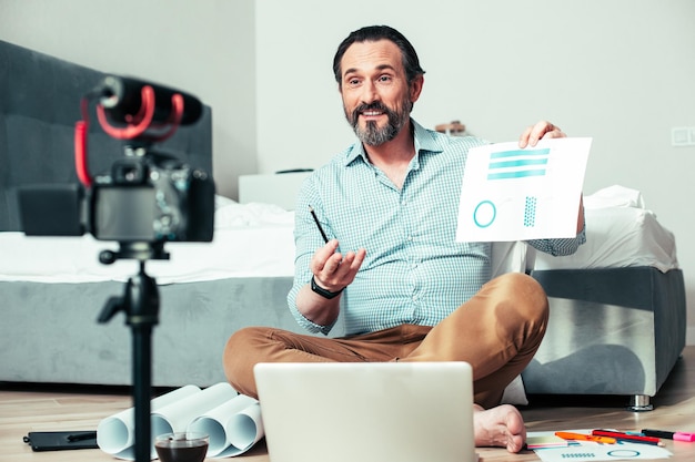 Confident smiling man looking at the camera on tripod while holding a sheet of paper with graphic charts Blueprints and laptop near him