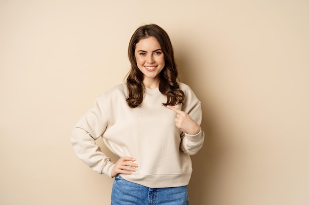 Confident smiling modern woman pointing finger at herself selfpromoting standing over beige background