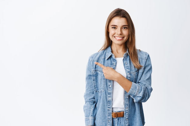 Confident smiling woman making announcement, advertising promo offer on copy space, pointing finger left and looking at front satisfied, white wall
