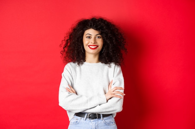 Fiduciosa donna sorridente con trucco e acconciatura riccia, braccia incrociate sul petto e dall'aspetto professionale, muro rosso