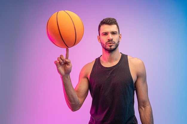 Confident sportsman spinning basketball ball on finger. Young bearded european basketball player looking at camera. Isolated on blue and pink background. Studio shoot. Copy space