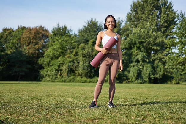 Confident and sporty young asian woman standing in park with\
rubber mat wearing sportswoman smiling