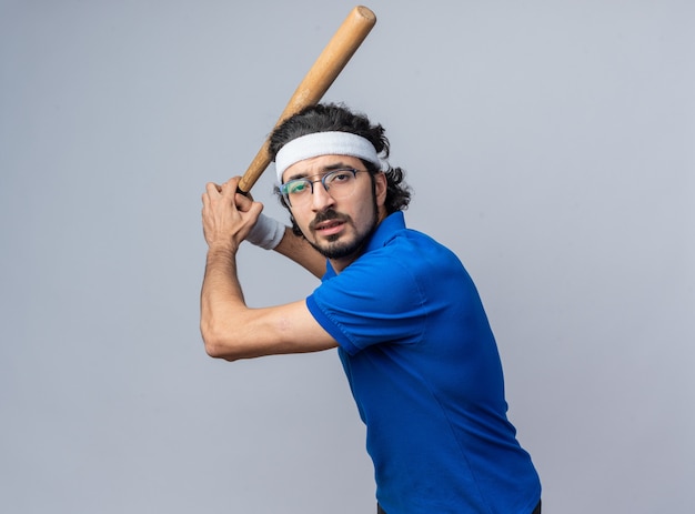 Confident standing in fighting pose young sporty man wearing headband with wristband holding baseball bat 