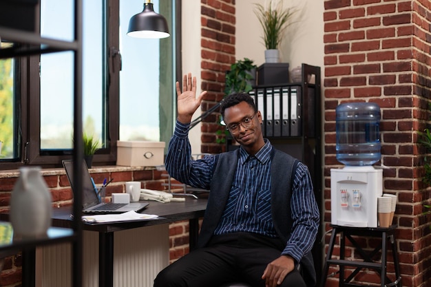 Confident startup employee sitting at desk with laptop waving hello in brick wall office. Small business owner with glasses smiling in front of portable computer doing hand gesture.