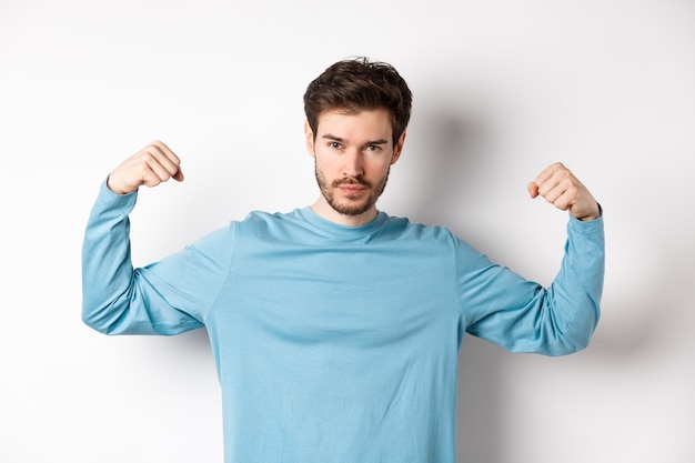 Confident and strong macho man flexing biceps, showing strength in muscles after gym workout, standing over white background