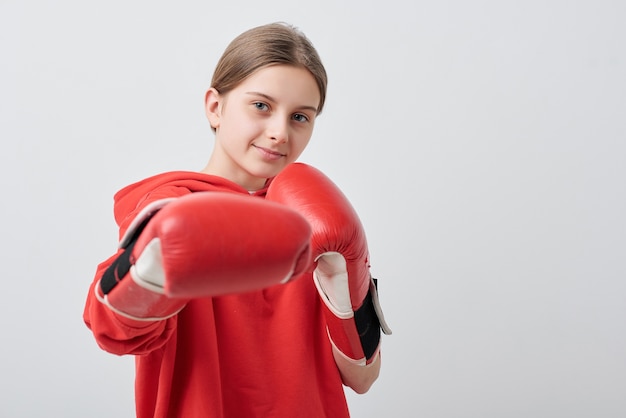 Fiducioso e forte ragazza adolescente in rosso activewear e guantoni da boxe facendo calcio mentre si esercita davanti alla telecamera in isolamento