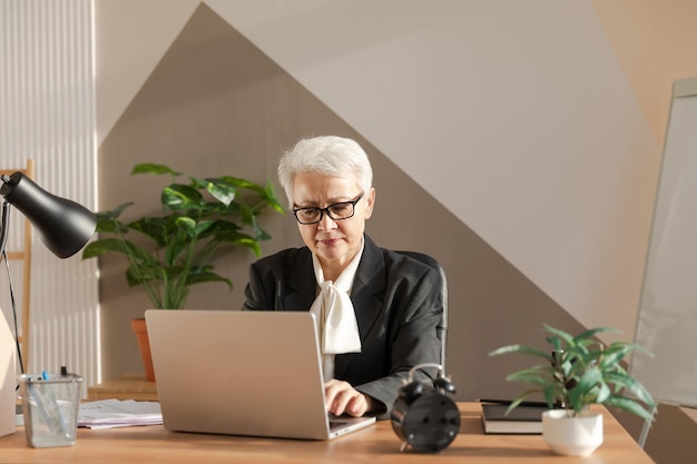 Confident stylish european middle aged senior woman using laptop at workplace Stylish older mature 60s gray haired lady businesswoman sitting at office table Boss leader teacher professional worker