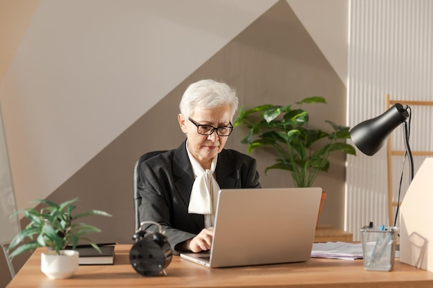 Confident stylish european middle aged senior woman using laptop at workplace Stylish older mature 60s gray haired lady businesswoman sitting at office table Boss leader teacher professional worker