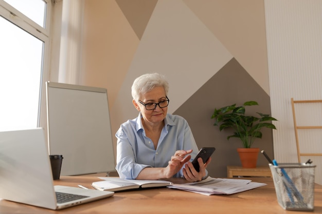 Confident stylish european middle aged senior woman using smartphone at workplace stylish older matu