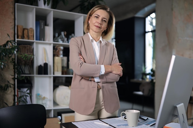 Confident stylish european middle aged woman standing at workplace, executive leader manager looking at camera.